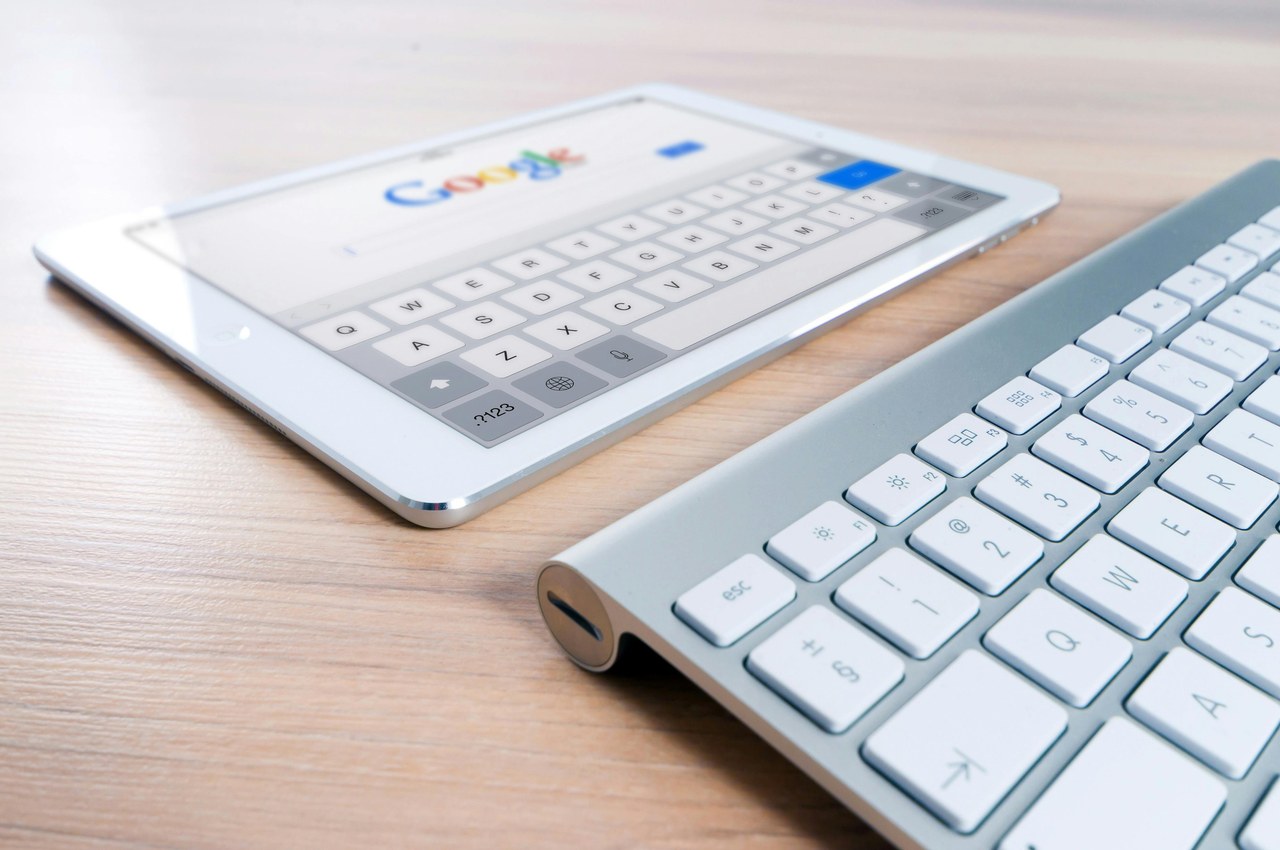 White tablet displaying Google homepage with on-screen keyboard beside aluminum desktop keyboard on wood surface, representing mobile-first design for local search optimization.
