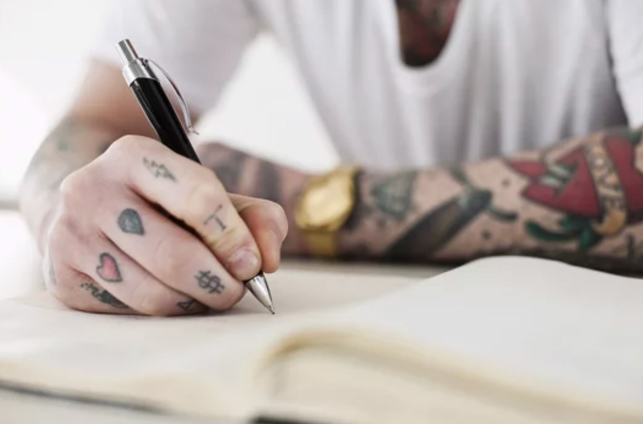 Close-up of a tattooed hand holding a pen over white paper, representing the personal expression and creativity in modern writing careers.