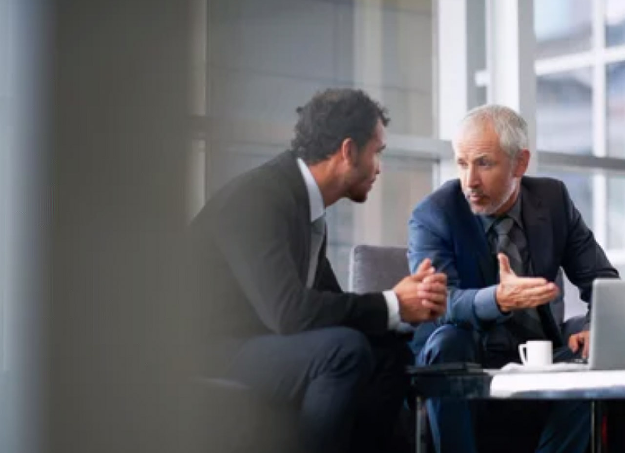 Two businessmen in suits have an animated conversation in a modern office, illustrating the shift toward conversational and peer-driven marketing approaches.