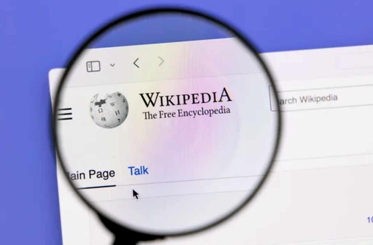 Magnifying glass enlarging Wikipedia homepage on computer screen, illustrating knowledge source research essential for entity SEO and building brand authority in zero-click search environments.