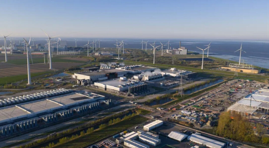 wind-farm-industrial-complex-aerial-coastal-renewable-energy Aerial view of a coastal industrial park with multiple wind turbines and warehouse facilities, illustrating renewable energy integration in commercial infrastructure for carbon footprint reduction.