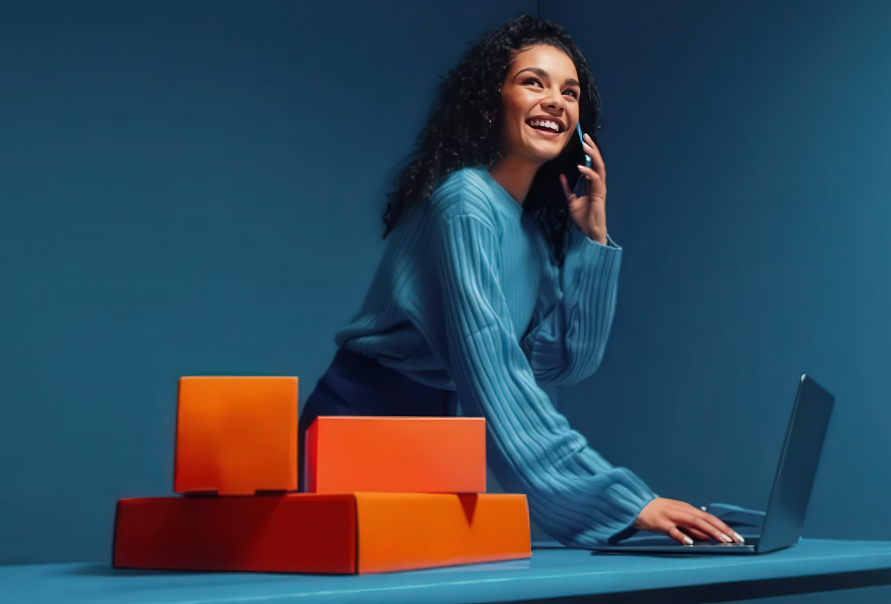 Woman wearing blue sweater smiles while working on laptop and speaking on phone beside product boxes, illustrating modern customer service multitasking in predictive support environments.