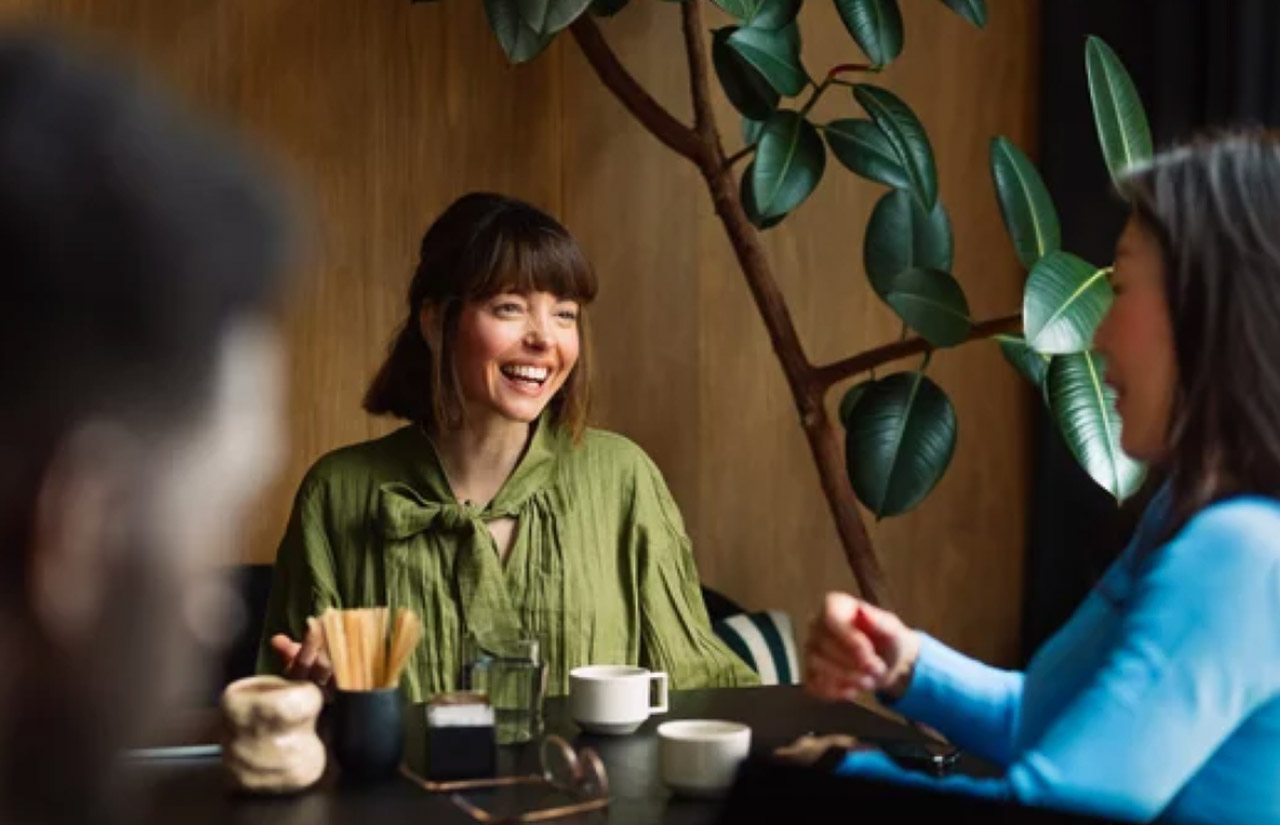 Woman in olive-green blouse laughing at cafe table during conversation, representing how memorable customer experiences generate word-of-mouth recommendations.
