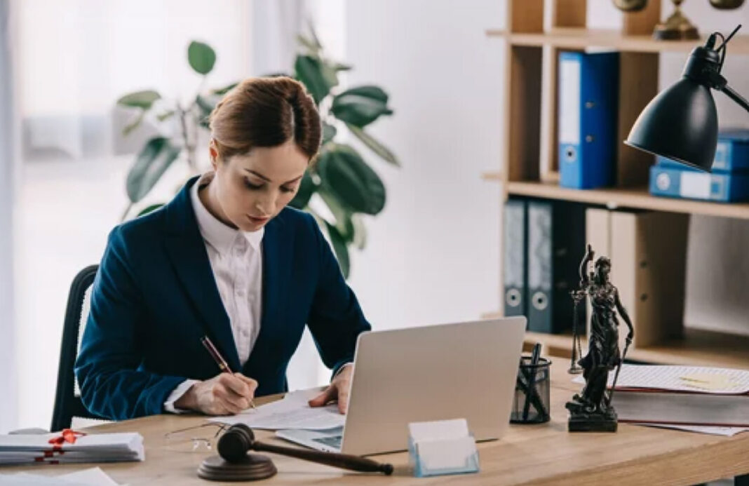 woman-lawyer-working-desk-laptop-legal-office-research-evaluation Professional woman in blazer works at desk with laptop and legal symbols, representing the research process for selecting law business directories.