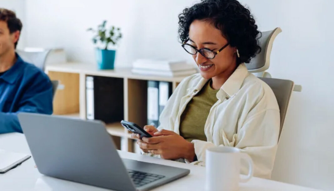 Professional uses smartphone and laptop at modern office desk, illustrating digital tools for online business discovery Woman seated at office desk uses smartphone while laptop is open beside her, representing modern digital tools for accessing online business directories.