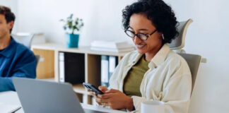 The Future of Online Directories Woman seated at office desk uses smartphone while laptop is open beside her, representing modern digital tools for accessing online business directories.