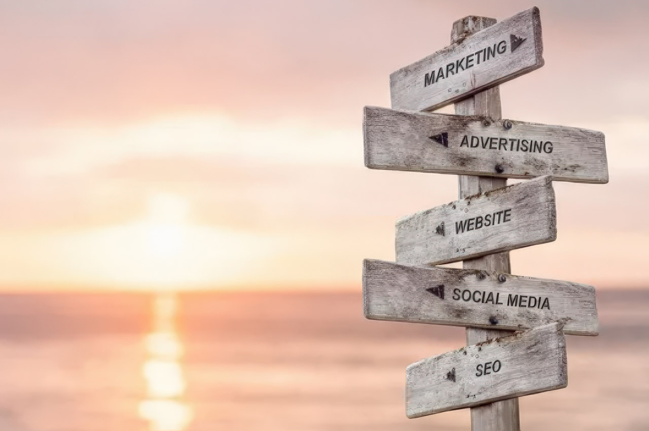 A weathered wooden directional signpost displays marketing, advertising, website, social media, and SEO signs against a sunset sky, representing strategic channels to enhance business listing visibility.